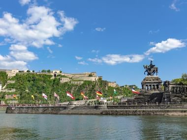 Blick auf die Festung Ehrenbreitstein am Hang und das Reiterstandbild am Deutschen Eck in Koblenz mit Rhein und bunten Flaggen.