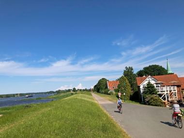 Two cyclists riding on paved path atop Elbe River dike near Lauenburg. Green grass slopes, river view left, traditional houses with church spire right.