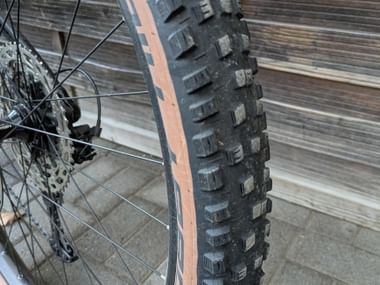 Close-up of a bicycle wheel with a knobby winter tire featuring deep tread blocks and beige sidewalls against a wooden fence background.