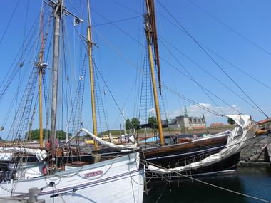 Traditional wooden sailing ships with tall masts moored in Helsingør harbor. Kronborg Castle with green copper roof visible in background under blue sky.