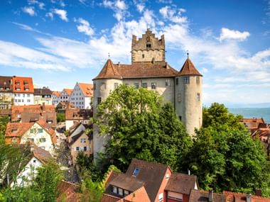 Mittelalterliche Alte Burg in Meersburg mit Steintürmen und roten Ziegeldächern, umgeben von bunten Häusern und grünen Bäumen mit Blick auf den Bodensee.
