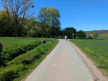 Paved bike path stretching through green fields with cyclists in the distance. Trees line the path under clear blue sky with hills in background.