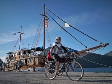 Radfahrer auf Hafenkai neben traditionellem Holzsegelschiff Panagiota mit zwei Masten und Takelage, Berge im Hintergrund sichtbar.