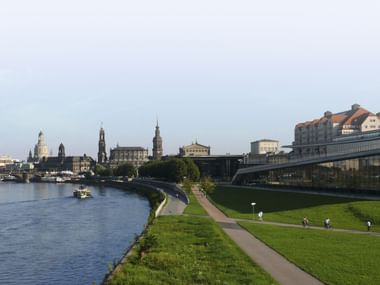 Blick auf Dresdens Elbufer mit dem Maritim Hotel Dresden, historischen Kirchtürmen und grüner Uferpromenade mit Fußgängern.
