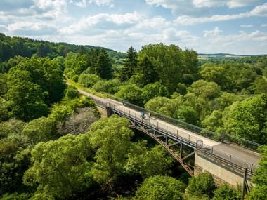 Aerial view of a curved concrete bridge on the Kyll cycle path near Stadtkyll, surrounded by lush green forest in the Eifel region.