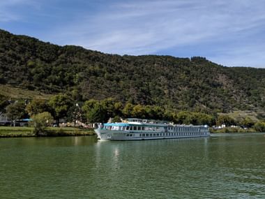MS Switzerland Flusskreuzfahrtschiff mit weißem Rumpf und blauem Oberdeck auf ruhigem grünem Wasser vor bewaldeten Hügeln und Dorf.
