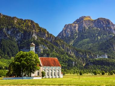 White church with red roof and bell tower in green meadow near Schwangau, with Neuschwanstein Castle visible on forested mountain slopes.