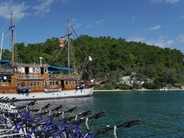 Traditionelles Holzschiff Panagiota im griechischen Hafen mit Reihen blauer Fahrräder am Kai. Klares türkisfarbenes Wasser und grüne Hügel.
