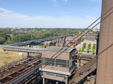 Blick vom Phoenix West Skywalk in Dortmund mit Industriebauten aus Backstein, Rohrleitungen und Hochofenresten vor grüner Landschaft.