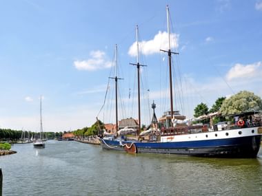 Historic three-masted sailing vessel with white hull and brown sails moored at a Dutch harbor jetty, with traditional buildings and marina in background.