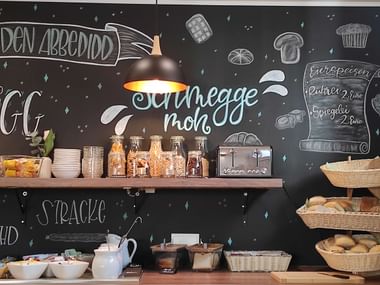 Frühstücksbuffet mit Kreidetafelwand, Holzregal mit Müsligläsern, Brotkörben und weißer Teekanne in der Pension Alte Scheune in Melsungen.