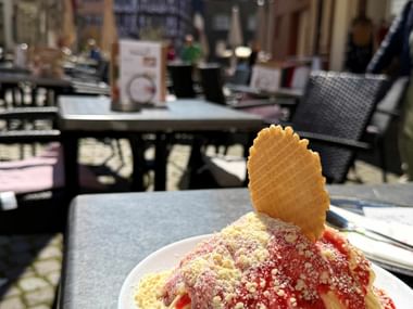 Spaghettieis mit Erdbeersoße und Waffel auf einer Café-Terrasse. Historische Gebäude und Außenbestuhlung im Hintergrund sichtbar.