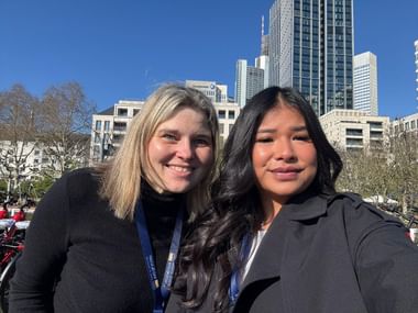 Zwei Frauen, Anja und Siri, lächeln in die Kamera mit modernen Wolkenkratzern und Bäumen im Hintergrund unter blauem Himmel.