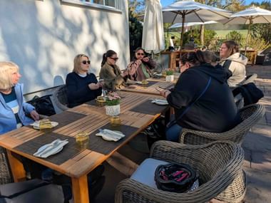 Group dining at wooden tables on sunny terrace at Waldschlösschen. White umbrellas provide shade, cyclists' gear visible on wicker chairs.