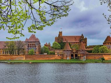 Malbork Castle viewed from across the river Malbork Castle complex with red brick walls and towers reflected in the river water, framed by green spring foliage under a cloudy sky.