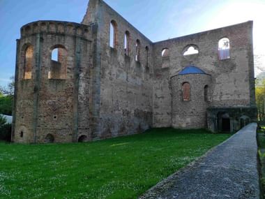 Mittelalterliche Kirchenruine mit hohen Backsteinmauern, Bogenfenstern und rundem Turm. Grüne Wiese im Vordergrund, gepflasterter Weg rechts.