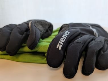 Two pairs of black padded winter cycling gloves with brand name visible, placed on a folded green jacket on a light wooden surface.