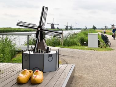 Small windmill model on wooden platform with yellow wooden shoes, cyclists on path, historic Kinderdijk windmills visible in background.