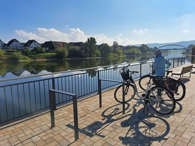 Radfahrer mit Fahrrad auf gepflasterter Weserpromenade in Beverungen, Blick auf ruhigen Fluss mit Häusern und Brücke im Hintergrund bei blauem Himmel.
