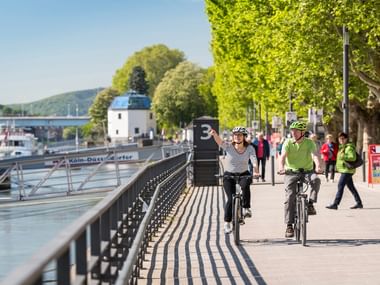Zwei Radfahrer fahren entlang eines gepflasterten Flussradwegs in Koblenz mit Flussschiffen, Brücke und grünen Bäumen an der Uferpromenade.