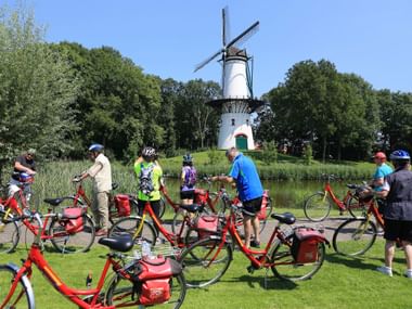 Group of cyclists with red rental bikes taking a break near a traditional white Dutch windmill in Tholen, surrounded by green trees.