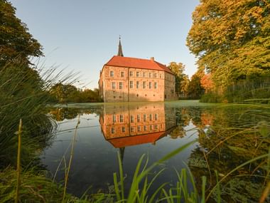 Historic Burg Lüdinghausen castle with red roof reflected in moat water, surrounded by autumn trees with golden foliage and reeds.