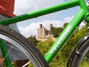 Mittelalterliche Ruinen der Burg Hanstein durch grünen Velociped-Fahrradrahmen sichtbar. Die Burg thront auf einem bewaldeten Hügel.