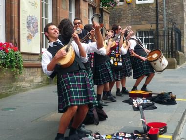 Group of Scottish street musicians performing in traditional green tartan kilts, playing mandolin, guitar and drums on a stone street.