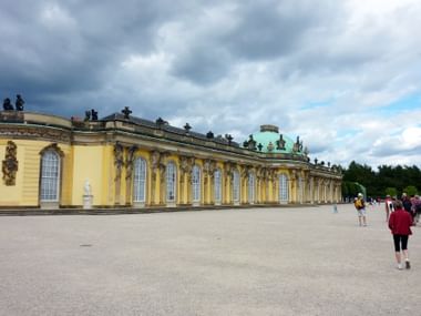 Gelbe Barockfassade von Schloss Sanssouci in Potsdam mit verzierten Fenstern und grüner Kupferkuppel, Besucher auf dem Hof.