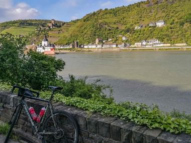 Black touring bicycle leaning against stone wall overlooking Rhine River. Hillside village with church and castle ruins visible across the water.
