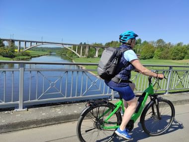 Radfahrer mit blauem Helm und Rucksack auf grünem Fahrrad auf Brücke über die Fulda. Große Bogenbrücke im Hintergrund unter klarem Himmel.