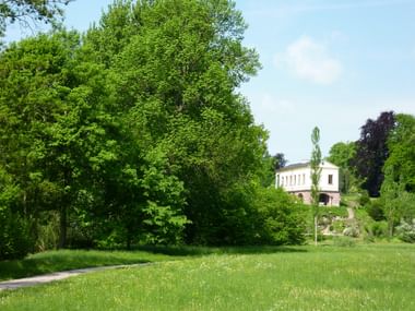 Park an der Ilm in Weimar mit grüner Wiese Grüne Wiese mit Spazierweg im Park an der Ilm, Weimar. Große Bäume rahmen ein weißes klassisches Gebäude im Hintergrund unter blauem Himmel.