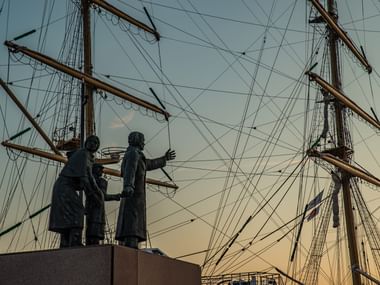 Emigrant Monument in Bremerhaven Bronze statue of two emigrants with luggage against historic sailing ship masts and rigging in Bremerhaven at sunset.