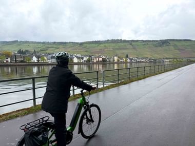 Radfahrer mit grünem Fahrrad und Helm auf Uferweg an der Mosel, blickt auf Dorf und Weinberge am gegenüberliegenden Ufer unter bewölktem Himmel.