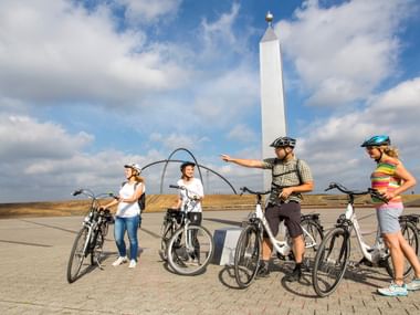 Vier Radfahrer mit Rädern stehen auf gepflastertem Platz an der Halde Hohenward. Weißer Obelisk und Metallbogen-Skulptur vor bewölktem Himmel.
