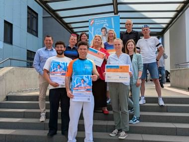 Group of people on stairs holding organ donation campaign posters in front of a modern building with glass canopy.