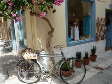 White bicycle with flower basket parked on stone street in Nafplion. Blue-trimmed shop displays white clothing, with bougainvillea overhead.