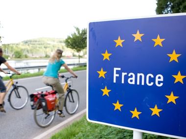 Blaues EU-Grenzschild mit 'France' und gelben Sternen, zwei Radfahrer mit Rädern unscharf im Hintergrund auf asphaltierter Straße.
