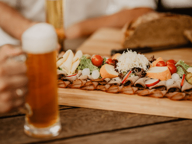 Glass of amber beer with foam next to wooden platter of Austrian snacks including cheese, nuts, vegetables, and bread in Mühlviertel style.