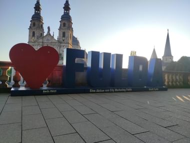 Großer 'I Love Fulda' Schriftzug mit rotem Herzsymbol auf einem Platz. Fuldaer Dom mit barocken Zwillingstürmen im Hintergrund unter klarem Himmel.