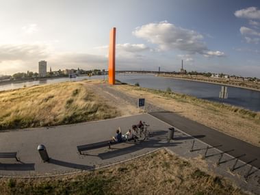 Orange Rheinorange Skulptur steht prominent auf grasbewachsenem Hügel mit Blick auf den Rhein in Duisburg mit Radfahrern auf Aussichtsplattform.
