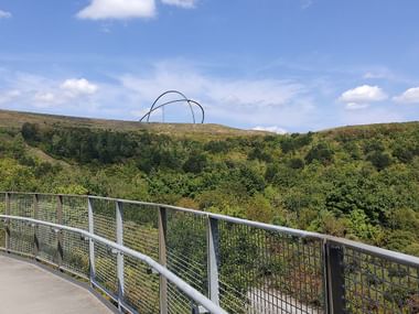 Gehweg mit Metallgeländer auf der Halde Hoheward mit Blick auf grüne Vegetation. Große Bogenskulptur auf dem Hügel gegen blauen Himmel.