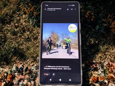 Smartphone displaying Velociped WhatsApp channel with photo of cyclists on path. Screen shows welcome message and Velociped logo on autumn leaves.