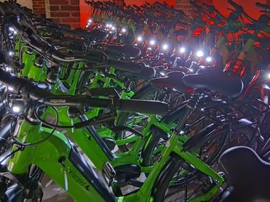Multiple rows of bicycles parked indoors, featuring bright green bikes in the foreground and darker bikes behind, against a red brick wall.