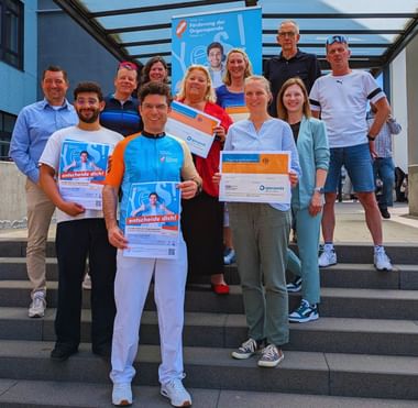 Group of people on steps holding organ donation certificates and banners. A blue banner reads 'Förderung der Organspende' in the background.
