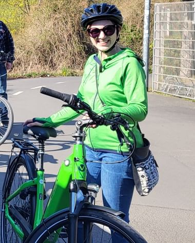 Christine smiling while standing with a green bicycle, wearing a green jacket, blue jeans, sunglasses, and a black cycling helmet on a bike path.