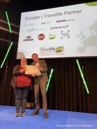 Two people on stage holding a Travelife Partner sustainability certificate. Behind them, a screen displays European travel partner logos.