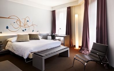 Modernes Doppelzimmer mit weißem Bett, dekorativer Wandkunst, lila Vorhängen, Stehlampe und schwarzen Lederstühlen im Hotel Continental Park in Luzern.