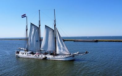 Two-masted sailing ship Elizabeth with white sails on calm blue water under clear sky, with coastal landscape in background.