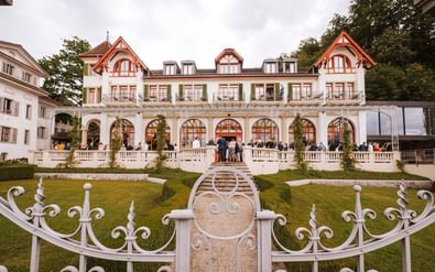 Hotel Seeburg in Luzern mit weißer Fassade, rot verzierten Giebeln, Bogenfenstern und Ziergarten mit weißem Dekorationszaun.